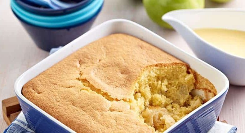 Golden-brown cake with a missing slice sits in a blue ceramic dish on a wooden board, next to a gravy boat filled with custard, bowls, and fresh green apples.