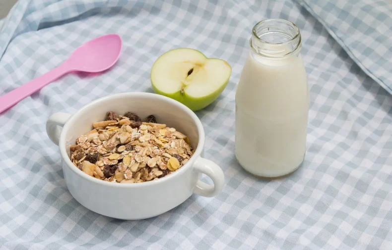 White bowl of muesli with glass bottle of milk, halved green apple, pink spoon on blue-checkered cloth background.